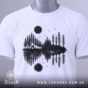 Футболка з природою, біла, складена на поверхні, фон світло-сірий, принт з чорними деревами, озером і сонцем, на футболці є етикетка з зеленим шнурком.
