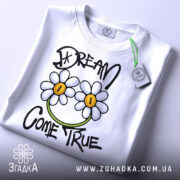 Футболка з ромашками, білого кольору, зображена на столі з легким ракурсом зверху, фон світло-сірий, принт з двома ромашками та написом 'DREAM COME TRUE' у чорному та зеленому кольорах, з етикеткою на зеленій стрічці.