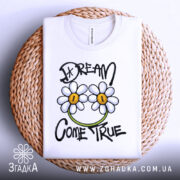 Футболка з ромашками, білого кольору, зображена на плетеній кошику, фон світло-сірий, принт з двома ромашками та написом 'DREAM COME TRUE' у чорному та зеленому кольорах.