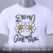 Футболка з ромашками, білого кольору, зображена на столі з легким ракурсом зверху, фон світло-сірий, принт з двома ромашками та написом 'DREAM COME TRUE' у чорному та зеленому кольорах, з етикеткою на зеленій стрічці.