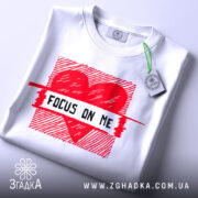 Футболка з серцем білого кольору, складена на поверхні, з верхнього ракурсу, фон світло-сірий, принт червоного кольору з написом 'FOCUS ON ME' та етикеткою з зеленим шнурком.