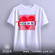 Футболка з серцем білого кольору, підвішена на вішалці, фон світло-сірий, принт червоного кольору з написом 'FOCUS ON ME' в центрі, вид спереду.