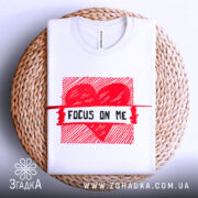 Футболка з серцем білого кольору, складена на плетеній кошику, з верхнього ракурсу, фон світло-сірий, принт червоного кольору з написом 'FOCUS ON ME'.