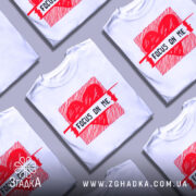 Футболка з серцем білого кольору, зображена на манекені з правого боку, фон світло-сірий, принт червоного кольору з написом 'FOCUS ON ME' в центрі.