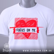 Футболка з серцем білого кольору, складена на поверхні, з верхнього ракурсу, фон світло-сірий, принт червоного кольору з написом 'FOCUS ON ME' та етикеткою з зеленим шнурком.