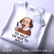 Футболка з шекспіром, білого кольору, зображена зверху, на столі, фон світлий, принт з карикатурним зображенням Шекспіра з пером в руці та написом 'TO MILF OR NOT TO MILF', на футболці є етикетка з зеленим шнурком.