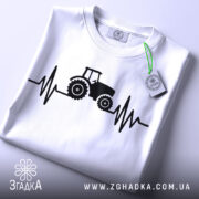 Футболка з трактором білого кольору, складена на поверхні, з видимим етикеткою, принт з трактором та лінією, що нагадує кардіограму, виконаний в чорному кольорі, фон світло-сірий.