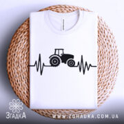Футболка з трактором білого кольору, складена на плетеній кошику, принт з трактором та лінією, що нагадує кардіограму, виконаний в чорному кольорі, фон світло-сірий.