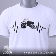 Футболка з трактором білого кольору, складена на поверхні, з видимим етикеткою, принт з трактором та лінією, що нагадує кардіограму, виконаний в чорному кольорі, фон світло-сірий.