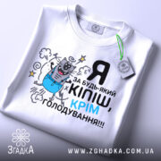 Футболка за будь-який кіпіш, біла, зображена зверху, на ній видно принт з котом у синіх штанах, текст чорний і блакитний, на комірці є етикетка.