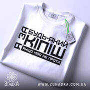 Футболка за кіпіш: біла футболка, складена на поверхні, з чорним принтом, що містить текст "Будь-який з Кіпіш, якщо моя не проти", вид зверху, фон світло-сірий.