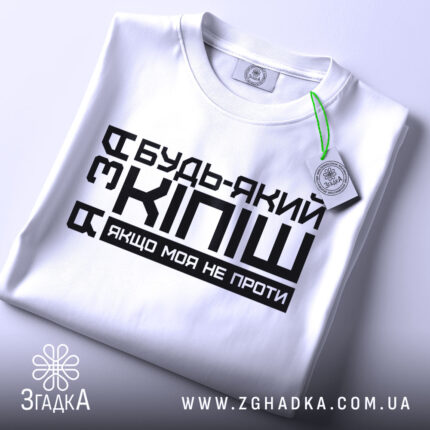 Футболка за кіпіш: біла футболка, складена на поверхні, з чорним принтом, що містить текст "Будь-який з Кіпіш, якщо моя не проти", вид зверху, фон світло-сірий.