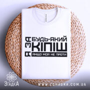 Футболка за кіпіш: біла футболка, складена на плетеній кошику, з чорним принтом, що містить текст "Будь-який з Кіпіш, якщо моя не проти", фон світло-сірий.