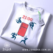 Футболка забіг до холодильника. Біла футболка, складена на поверхні, з принтом червоного холодильника та фігурами людей, що стрибають. Ракурс зверху, фон світлий, на футболці є етикетка з зеленим шнурком.