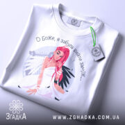 Футболка забула вийти заміж. Біла футболка, складена на столі, з видимим принтом дівчини з рожевим волоссям. На футболці видно етикетку з зеленим шнурком. Фон світлий, що підкреслює яскравість принта.