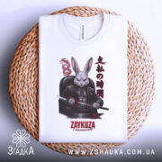 Футболка заяча мафія, біла, зображена зверху, складена на плетеній кошику, фон світлий, принт з зображенням кролика в чорному костюмі з мечем, текст на принті японською мовою та написом 'ZAYKUZA'.