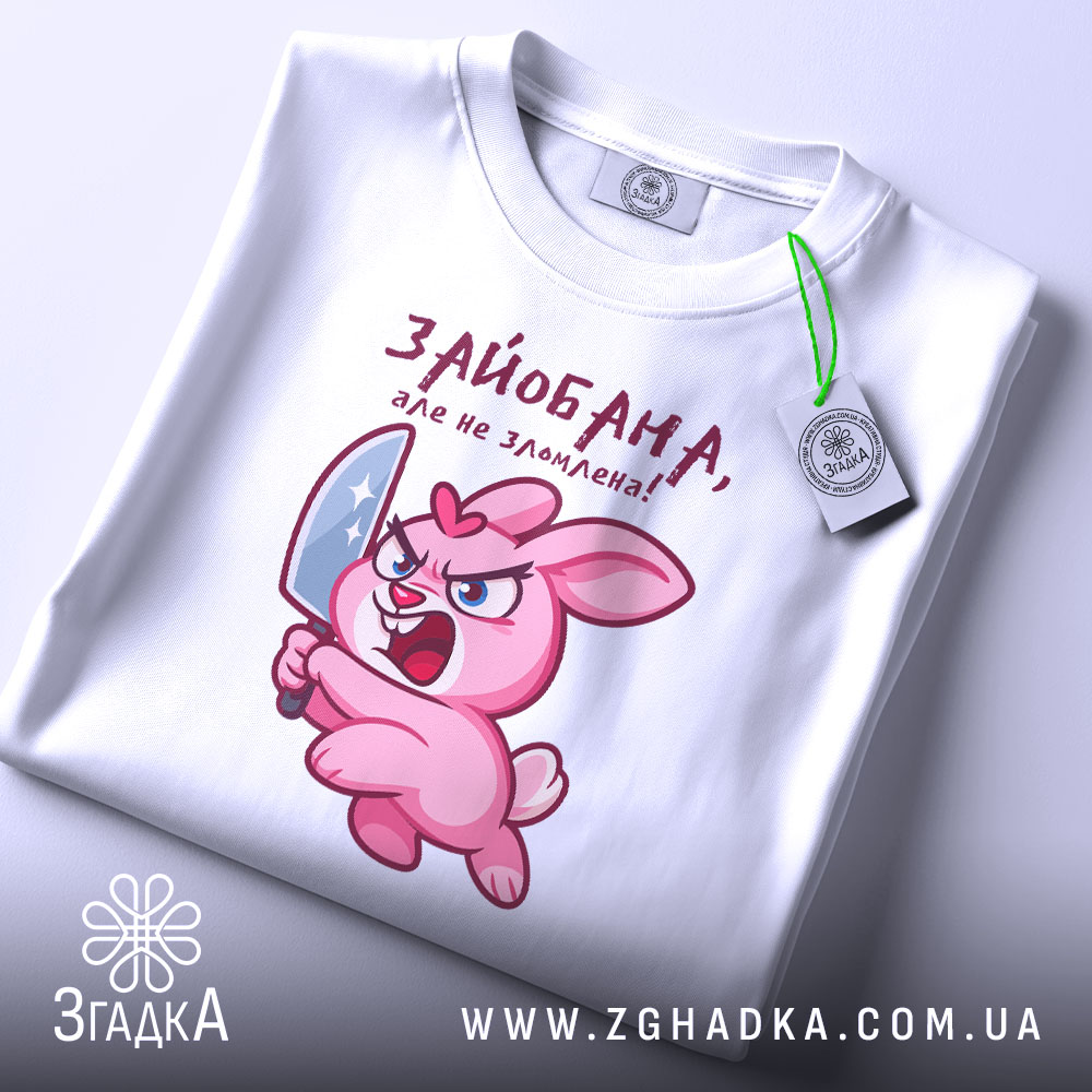 Футболка зайобана але не зломлена, біла, складена на поверхні, фон світло-сірий, принт з рожевим зайцем, що тримає ніж, з виразом гніву, з етикеткою на шнурку.