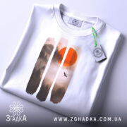 Футболка захід сонця, біла, складена на поверхні, фон світлий, принт з червоним сонцем та лісом в тумані, на футболці є етикетка з зеленим шнурком.