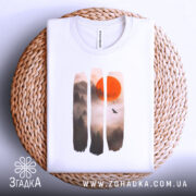 Футболка захід сонця, біла, складена на плетеній кошику, фон світлий, принт з червоним сонцем та лісом в тумані, футболка акуратно розкладена.