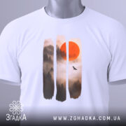 Футболка захід сонця, біла, складена на поверхні, фон світлий, принт з червоним сонцем та лісом в тумані, на футболці є етикетка з зеленим шнурком.