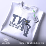 Футболка заходь коли що, біла, зображена зверху, складена, фон світло-сірий, принт з сірим котом з жовтими очима та текстом "ТИ заходь коли що...", на футболці є етикетка з зеленим шнурком.