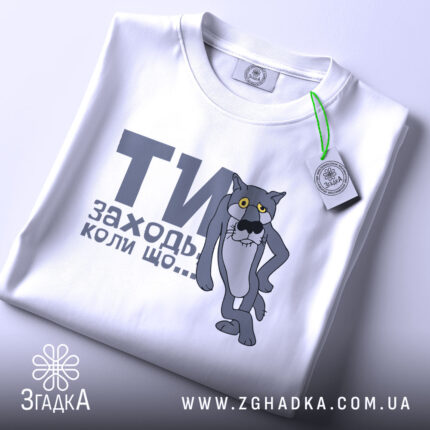 Футболка заходь коли що, біла, зображена зверху, складена, фон світло-сірий, принт з сірим котом з жовтими очима та текстом "ТИ заходь коли що...", на футболці є етикетка з зеленим шнурком.