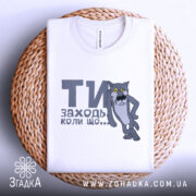 Футболка заходь коли що, біла, зображена зверху, складена на плетеній основі, фон білий, принт з сірим котом з жовтими очима та текстом "ТИ заходь коли що...".