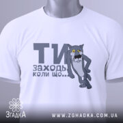 Футболка заходь коли що, біла, зображена зверху, складена, фон світло-сірий, принт з сірим котом з жовтими очима та текстом "ТИ заходь коли що...", на футболці є етикетка з зеленим шнурком.