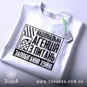 Футболка запобігання хуйні. Біла футболка, складена на поверхні, з видимим принтом на грудях. Принт чорного кольору з текстом "Національна агенція з питань запобігання хуйні". На футболці є етикетка з зеленим шнурком. Фон світло-сірий.