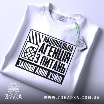 Футболка запобігання хуйні. Біла футболка, складена на поверхні, з видимим принтом на грудях. Принт чорного кольору з текстом "Національна агенція з питань запобігання хуйні". На футболці є етикетка з зеленим шнурком. Фон світло-сірий.