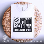 Футболка запобігання хуйні. Біла футболка, складена на плетеній основі, з видимим принтом на грудях. Принт чорного кольору з текстом "Національна агенція з питань запобігання хуйні". Фон світло-сірий, що створює контраст з текстурою основи.