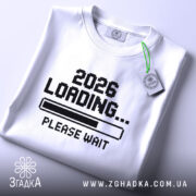 Футболка завантаження року, складена біла футболка з принтом '2026 LOADING... PLEASE WAIT' у чорному кольорі, на фоні світло-сірого кольору, з етикеткою зеленого кольору.