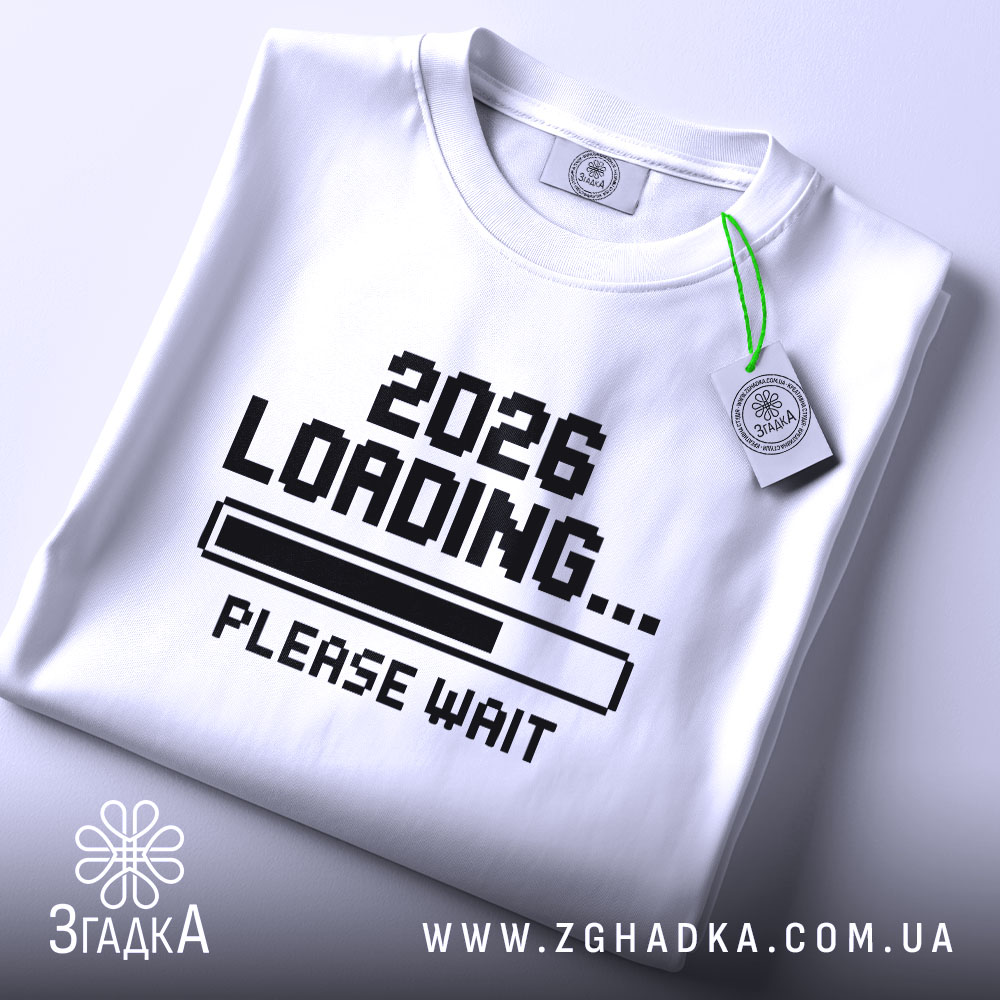 Футболка завантаження року, складена біла футболка з принтом '2026 LOADING... PLEASE WAIT' у чорному кольорі, на фоні світло-сірого кольору, з етикеткою зеленого кольору.