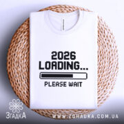 Футболка завантаження року, складена біла футболка з принтом '2026 LOADING... PLEASE WAIT' у чорному кольорі, на фоні плетеного кошика, деталі принта чіткі.