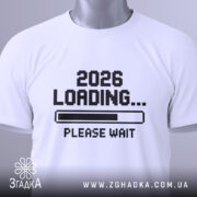 Футболка завантаження року, складена біла футболка з принтом '2026 LOADING... PLEASE WAIT' у чорному кольорі, на фоні світло-сірого кольору, з етикеткою зеленого кольору.
