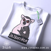 Футболка зазбогійся, білого кольору, зображена зверху, на ній видно принт з ведмедем, що має червоні очі, та напис 'ЗАБОТИСЯ', на футболці також є етикетка з зеленим шнурком.