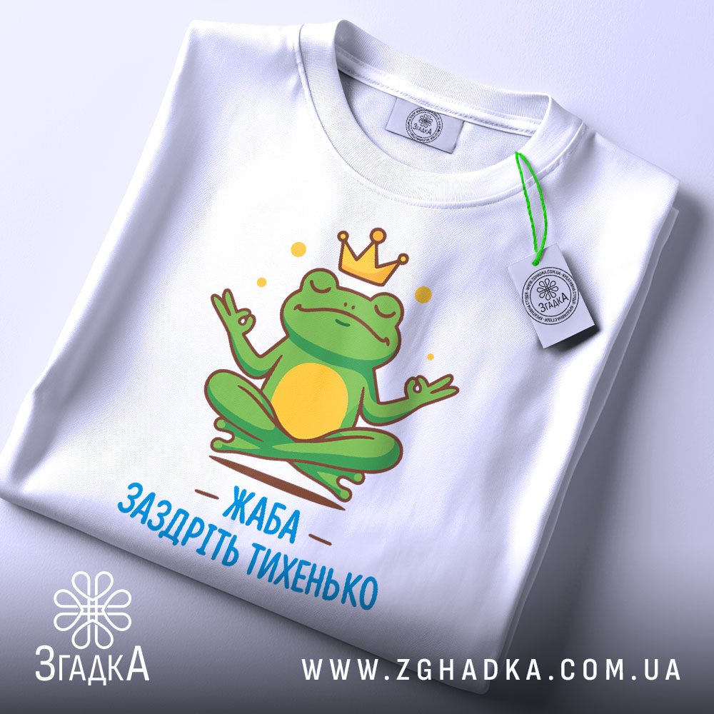 Футболка заздріть тихенько: біла футболка з малюнком жаби, що сидить у медитаційній позі, з зеленим тегом на комірці, принт яскравий, з короною на голові жаби та написом 'ЖАБА ЗАЗДРІТЬ ТИХЕНЬКО'.