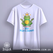 Футболка заздріть тихенько: біла футболка, підвішена на вішалці, з малюнком жаби в медитаційній позі, принт яскравий, з короною на голові жаби та написом 'ЖАБА ЗАЗДРІТЬ ТИХЕНЬКО'.