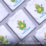 Футболка заздріть тихенько: біла футболка з малюнком жаби, що сидить у медитаційній позі, на фоні світло-сірого градієнту, принт яскравий, з короною на голові жаби та написом 'ЖАБА ЗАЗДРІТЬ ТИХЕНЬКО'.