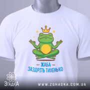 Футболка заздріть тихенько: біла футболка з малюнком жаби, що сидить у медитаційній позі, з зеленим тегом на комірці, принт яскравий, з короною на голові жаби та написом 'ЖАБА ЗАЗДРІТЬ ТИХЕНЬКО'.