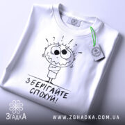 Футболка зберігайте спохуй. Біла футболка, складена на столі, з видимим ярликом, фон світлий, принт з мультяшним персонажем, що виглядає здивованим, з написом 'Зберігайте спохуй!' під ним.