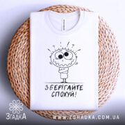 Футболка зберігайте спохуй. Біла футболка, складена на плетеному кошику, фон світлий, принт з мультяшним персонажем, що виглядає здивованим, з написом 'Зберігайте спохуй!' під ним.