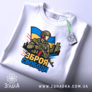 Футболка зброя свободи, біла, зображена на столі, фон світло-сірий, принт з військовим, що тримає зброю, на фоні українського прапора, з етикеткою на рукаві.