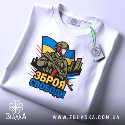 Футболка зброя свободи, біла, зображена на столі, фон світло-сірий, принт з військовим, що тримає зброю, на фоні українського прапора, з етикеткою на рукаві.
