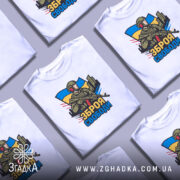 Футболка зброя свободи, біла, зображена на манекені, фон світло-сірий, принт з військовим, що тримає зброю, на фоні українського прапора та міського пейзажу.