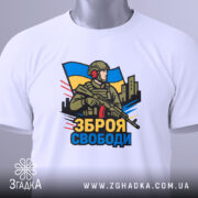 Футболка зброя свободи, біла, зображена на столі, фон світло-сірий, принт з військовим, що тримає зброю, на фоні українського прапора, з етикеткою на рукаві.