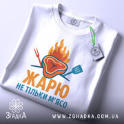Футболка жарю м`ясо, біла, зображена з верхнього ракурсу, фон світло-сірий, принт з зображенням шматка м'яса з вогнем і кухонними приладами, текст "ЖАРЮ НЕ ТІЛЬКИ М'ЯСО" яскравими кольорами, на футболці є етикетка.