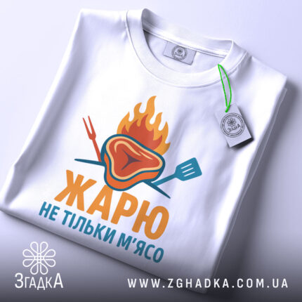 Футболка жарю м`ясо, біла, зображена з верхнього ракурсу, фон світло-сірий, принт з зображенням шматка м'яса з вогнем і кухонними приладами, текст "ЖАРЮ НЕ ТІЛЬКИ М'ЯСО" яскравими кольорами, на футболці є етикетка.
