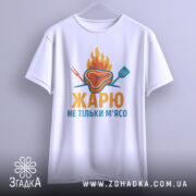 Футболка жарю м`ясо, біла, зображена з фронтального ракурсу, фон світло-сірий, принт з зображенням шматка м'яса з вогнем і кухонними приладами, текст "ЖАРЮ НЕ ТІЛЬКИ М'ЯСО" яскравими кольорами, футболка підвішена на вішалці.