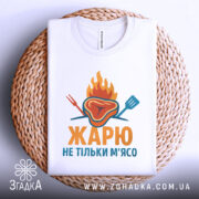 Футболка жарю м`ясо, біла, зображена з верхнього ракурсу, фон світло-сірий, принт з зображенням шматка м'яса з вогнем і кухонними приладами, текст "ЖАРЮ НЕ ТІЛЬКИ М'ЯСО" яскравими кольорами, футболка складена на плетеній основі.